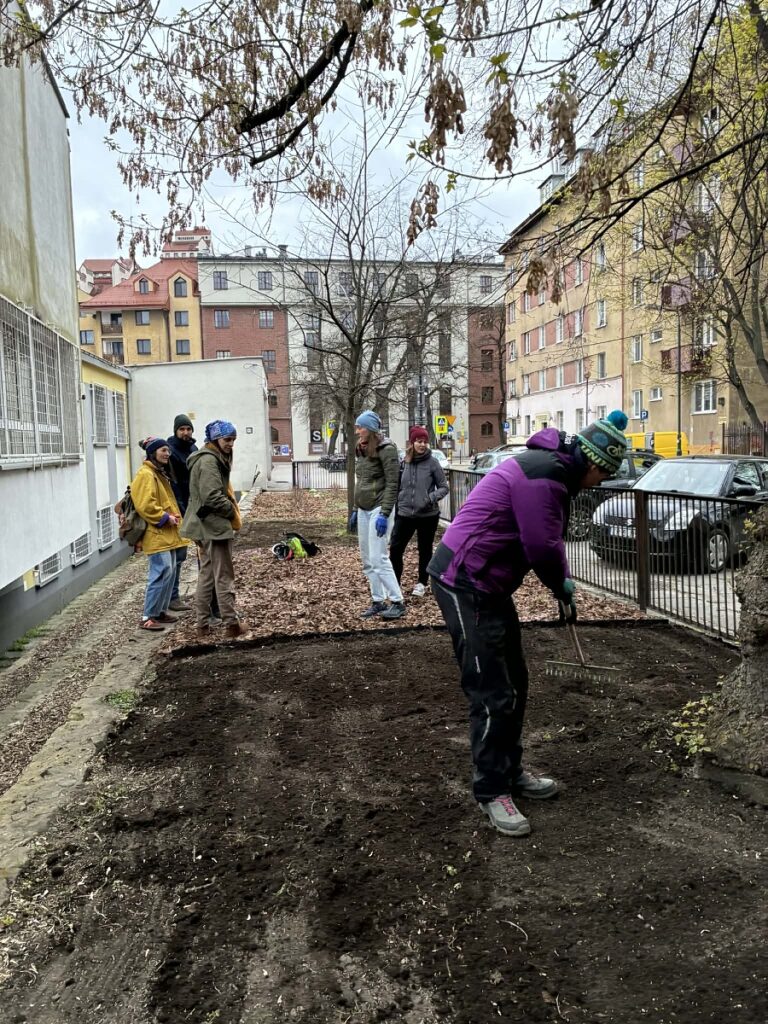 końcówka prac w ogródku, jedna osoba grabi ziemię, którą przykryto nasiona trawy, a reszta rozmawia